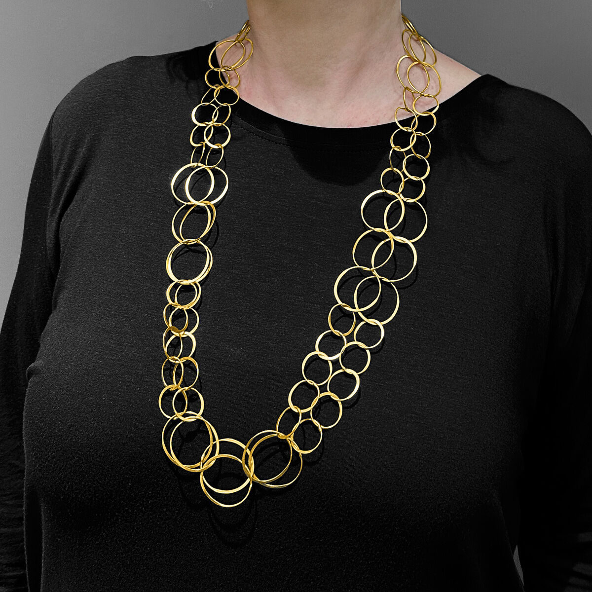 Frau mit blonder Frisur trägt eine lange Halskette mit verbundenen goldenen Ringen aus vergoldetem Silber über einem schwarzen Oberteil. Grau-weißer Hintergrund.