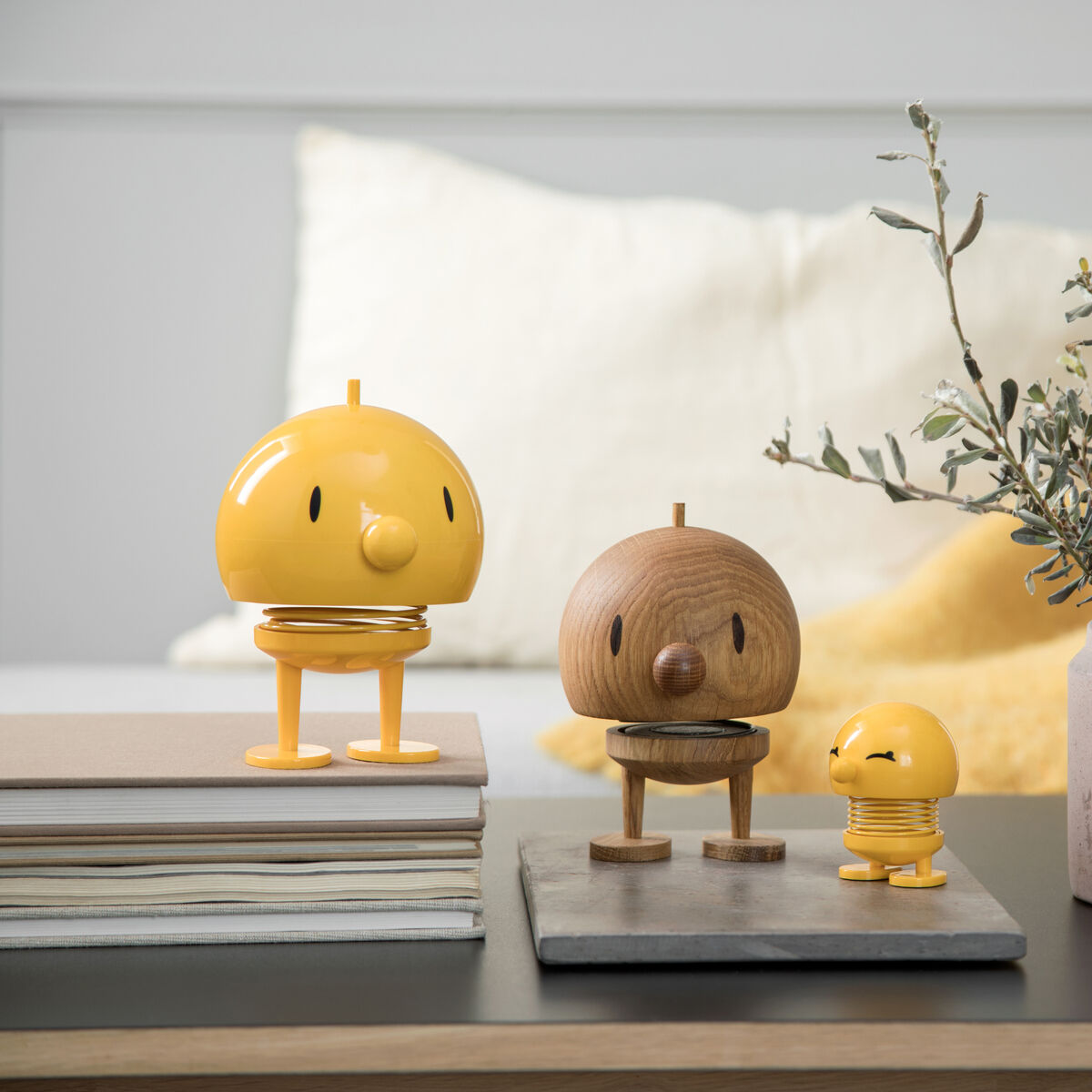 Bild von drei Hoptimist Bumble »Eiche« Figuren, die in einem modernen Wohnzimmer auf Büchern und Steinplatten platziert sind. Die mittlere Figur ist aus hellem Holz gefertigt, während die anderen beiden in verschiedenen Gelbtönen leuchten.