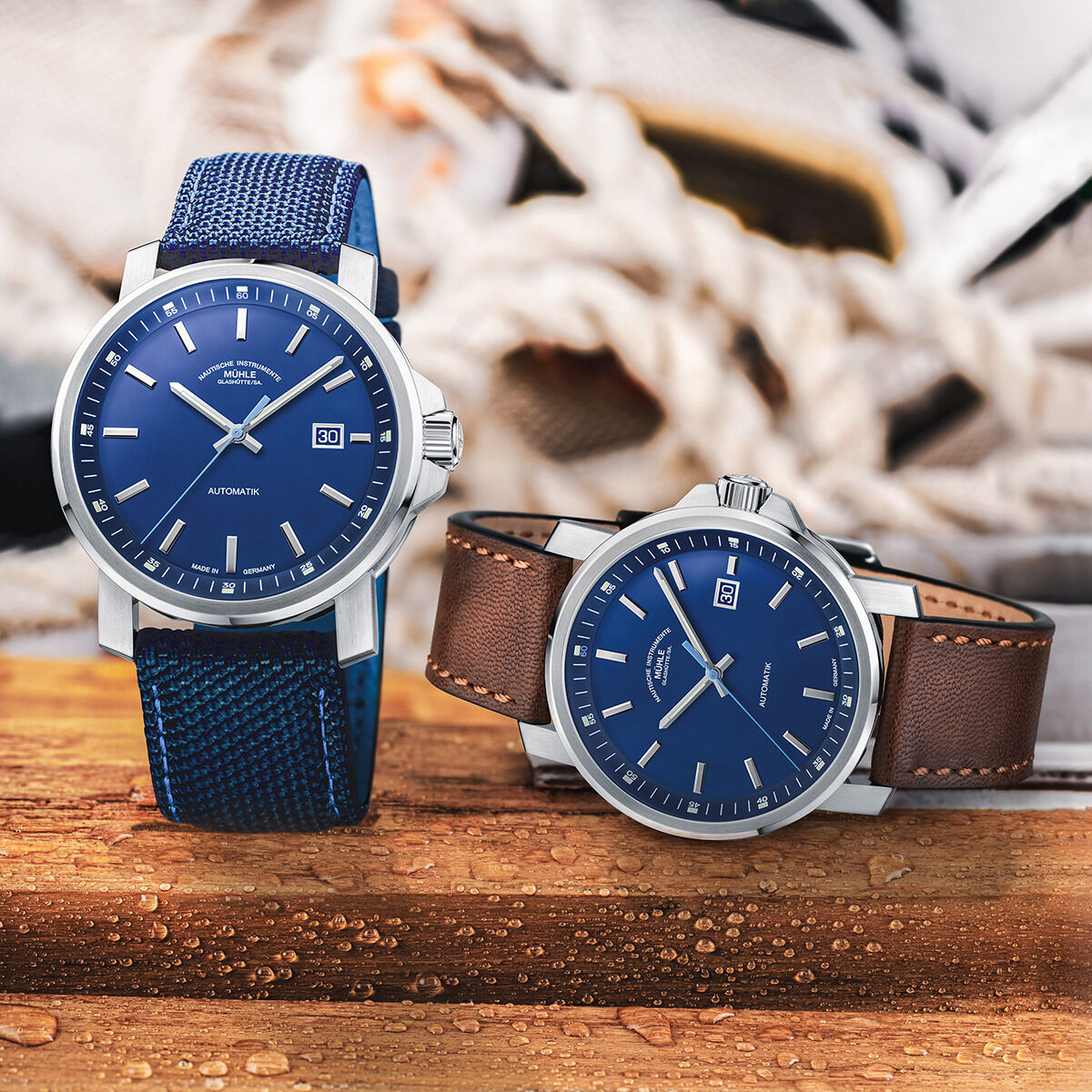 Zwei Armbanduhren der ZEIT-Sonderedition »29er Big Deep Blue« von Mühle-Glashütte auf einem Holzuntergrund. Eine Uhr hat ein blaues Textilarmband, die andere ein braunes Lederarmband. Im Hintergrund sind unscharfe maritime Elemente und Wassertropfen.