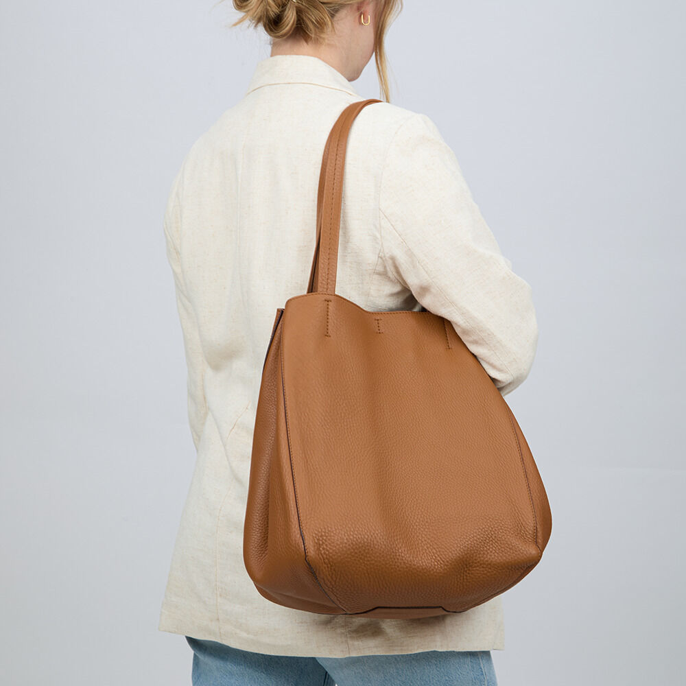 Frau mit heller Jacke und blauer Jeans trägt eine große, cognacfarbene Ledertasche „ZEIT-Tasche »Cep«“ von Guerer & Guerer über der Schulter. Die Tasche wirkt hochwertig, schlicht, mit langen Henkeln und weichem, natürlich strukturiertem Leder.