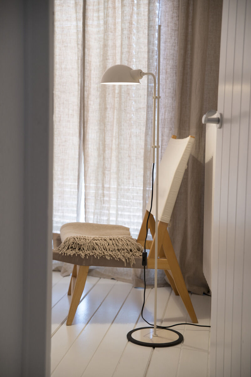 Eine moderne Stehleuchte »Funiculi« in einem minimalistischen Wohnzimmer. Die Lampe ist weiß und steht neben einem Stuhl aus Holz mit einem beigen Sitzkissen. Helle Vorhänge und ein Holzfußboden sind ebenfalls zu sehen.