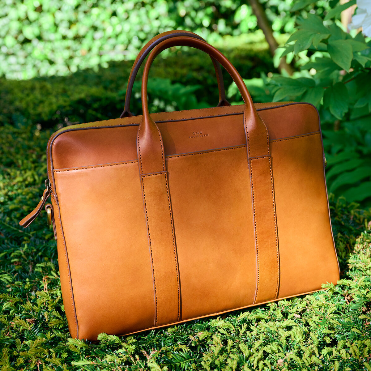 Eine braune Ledertasche »Harvey« steht auf grüner Vegetation, umgeben von Pflanzen im Hintergrund. Die Tasche hat zwei robuste Griffe und ein elegantes Design.
