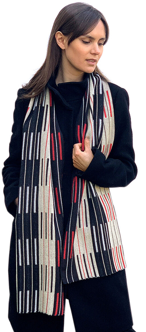 Eine Frau trägt einen eleganten Seidenschal namens »Doublé« mit einem minimalistischen Streifenmuster in Schwarz, Weiß und Rot. Sie trägt einen dunklen Mantel und scheint sich wohlzufühlen.