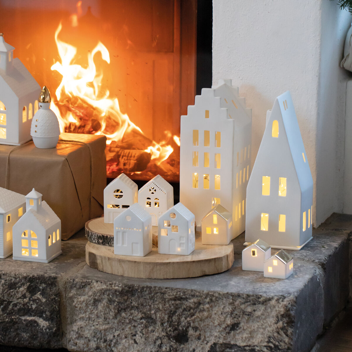 Mehrere beleuchtete weiße Keramikhäuser stehen dekorativ auf einem Kamin, vor einem brennenden Feuer, und schaffen eine gemütliche Weihnachtsatmosphäre.