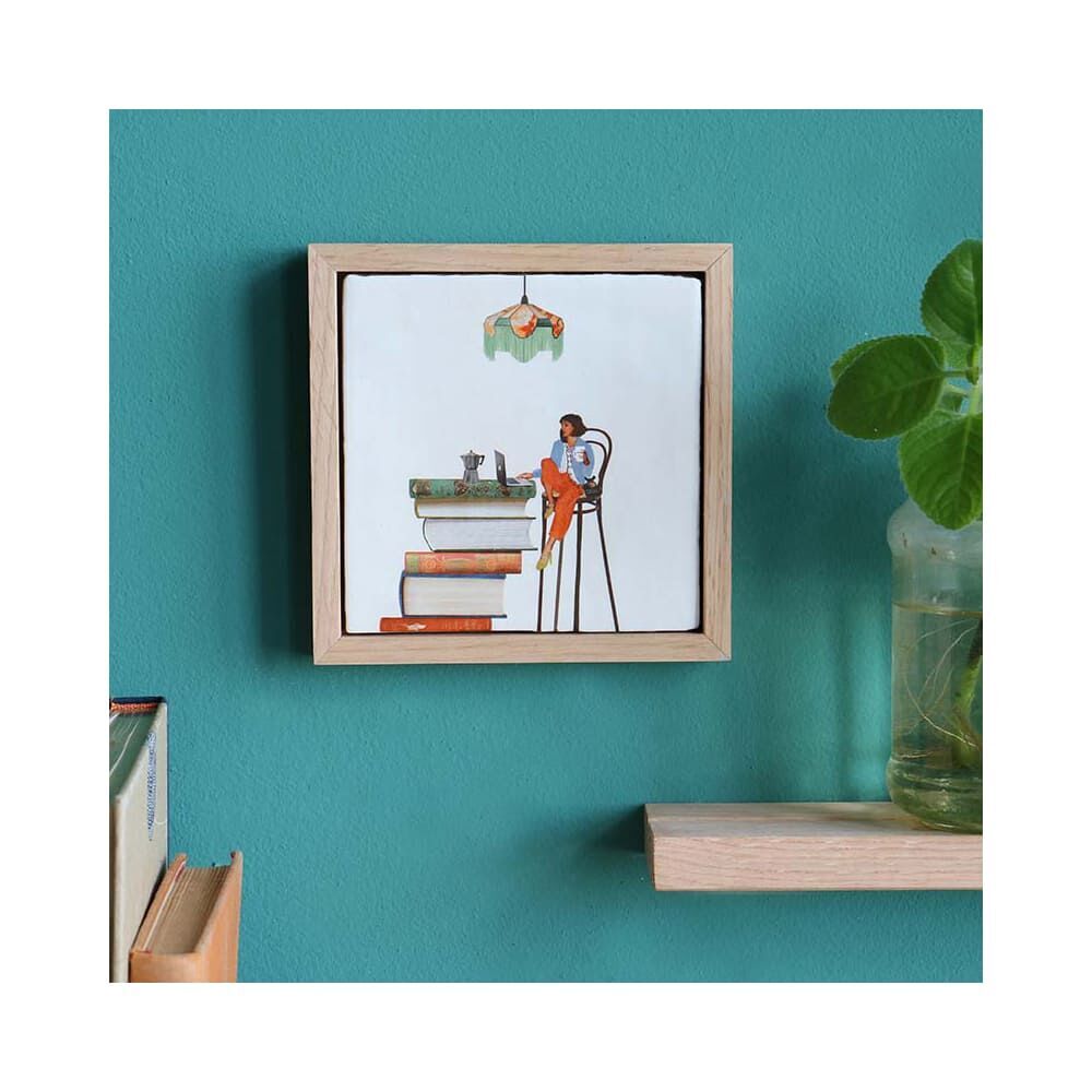 Quadratische Keramikfliese mit Holzbilderrahmen an einer türkisfarbenen Wand. Die Fliese zeigt eine humorvolle Illustration einer Person, die auf einem hohen Stuhl an einem Schreibtisch sitzt, gestapelte Bücher als Unterlage nutzt und von einer hängenden Lampe beleuchtet wird. Rechts neben der Fliese steht eine Glasvase mit grüner Pflanze auf einem Holzregal.
