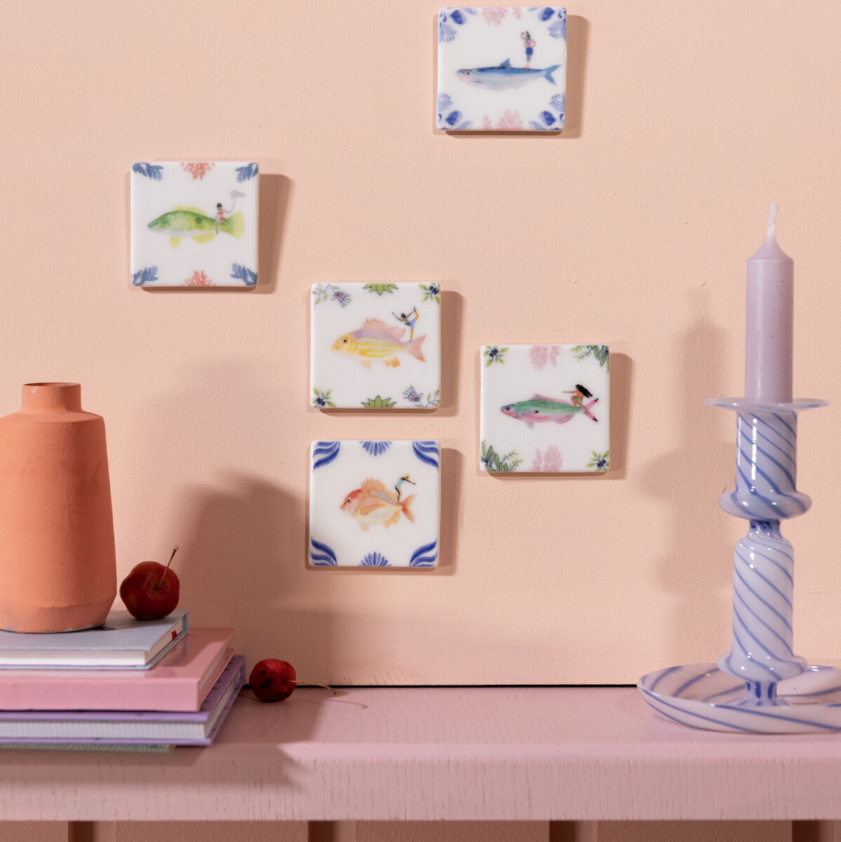 Ein Arrangement von fünf quadratischen Keramikfliesen mit verschiedenen Tiermotiven an einer pastellfarbenen Wand. Auf einem Regal darunter befindet sich eine Terrakottavase neben gestapelten Büchern und einer Kerze in einem gestreiften Kerzenhalter.