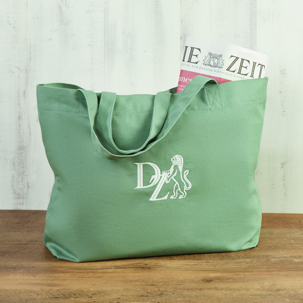 Eine grüne Canvas-Tasche mit dem aufgedruckten Logo der ZEIT in Weiß. Sie enthält eine Zeitung und steht auf einer Holztischplatte vor einem weiß gestrichenen Hintergrund.
