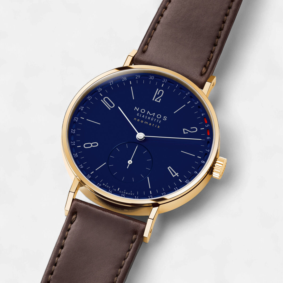 Elegante Armbanduhr NOMOS ZEIT-Sonderedition Tangente Gold neomatik 38 Update mit 18-karätigem Goldgehäuse, dunkelblauem emaillelackierten Zifferblatt, silbernen Ziffern, roten Datumsmarkierungen und dunkelbraunem Lederarmband auf hellem Hintergrund.