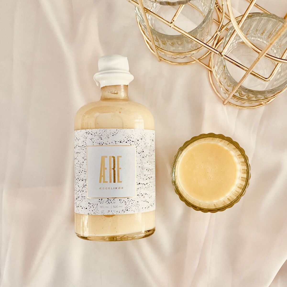 Eine Flasche Aere Eierlikör liegt auf einem beigen Tuch neben einem vollen Likörglas und einem goldenen, dekorativen Glasbehälter.