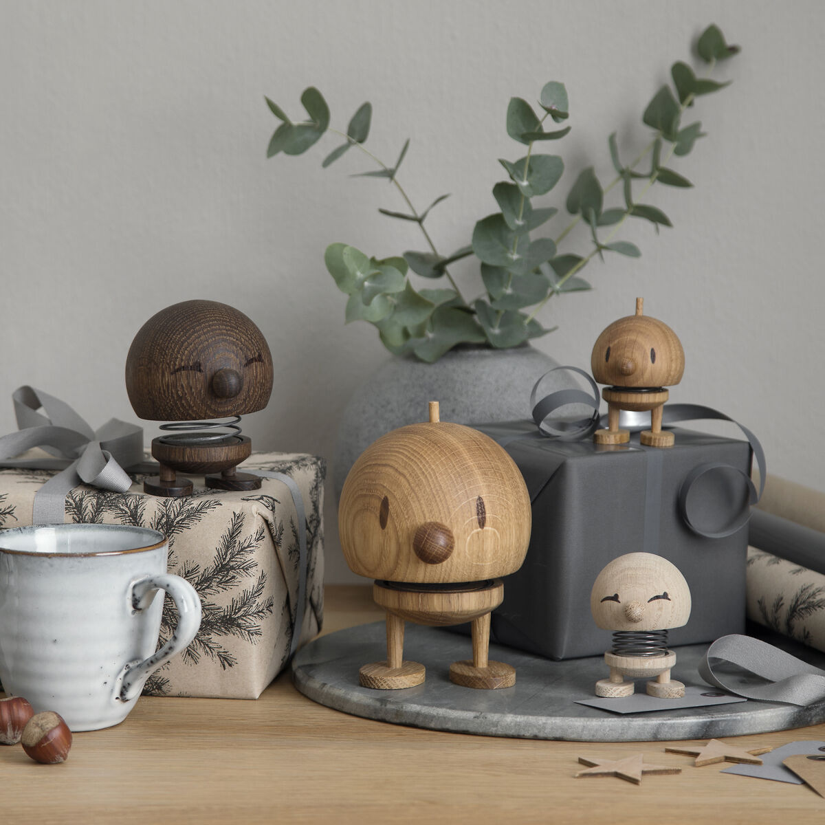 Eine Sammlung von Hoptimist Bumble Figuren in verschiedenen Brauntönen der Eiche steht auf einem Tisch. Die Szene ist minimalistisch und enthält Geschenkpakete, eine Tasse, Walnüsse und Eukalyptuszweige. Die Dekoration verleiht einen skandinavischen, natürlichen Flair.