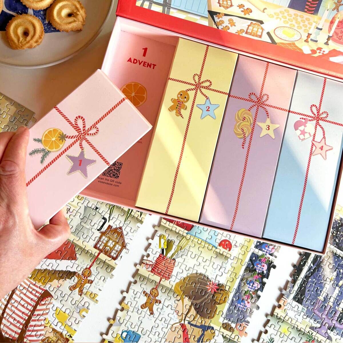 Abgebildet ist der Puzzle-Adventskalender »Sweet Christmas« von VISSEVASSE mit aufklappbarer Box, darin 24 nummerierte, bunte Fächer. Im Motiv eine weihnachtliche Küchenillustration. Unten Teile eines detailreichen Weihnachts-Puzzles, links ein Teller mit Keksen.