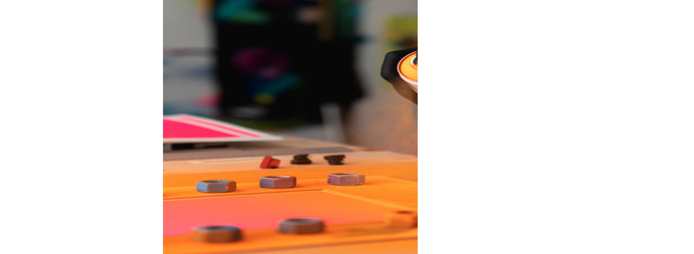 Detailaufnahme eines Künstlers, der mit schwarzer Handschuhen orangene Sprühfarbe auf Karton sprüht. Vor ihm liegen Werkzeuge und kleine bunte Objekte, im Hintergrund ist leicht verschwommen weiteres Material zu erkennen. Atmosphäre: kreativ und modern.