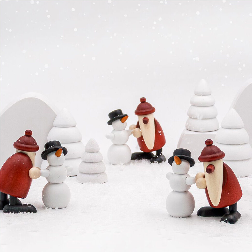 Drei handgefertigte Holzfiguren-Sets, jeweils bestehend aus einem Miniatur Weihnachtsmann und einem Schneemann mit schwarzem Hut, auf einer Schneelandschaft mit stilisierten weißen Winterbäumen. Die Figuren stammen von Björn Köhler.