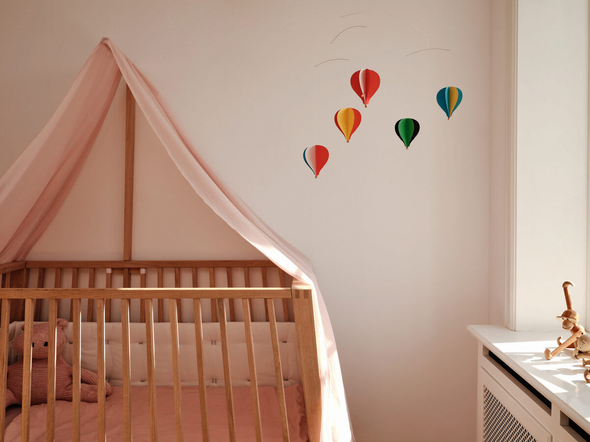 Ein Kinderzimmer mit einem hölzernen Kinderbett und rosa Bettwäsche. Über dem Bett ist eine Wanddekoration mit fünf bunten Heißluftballons in den Farben Rot, Gelb, Grün, Blau und Rosa angebracht.