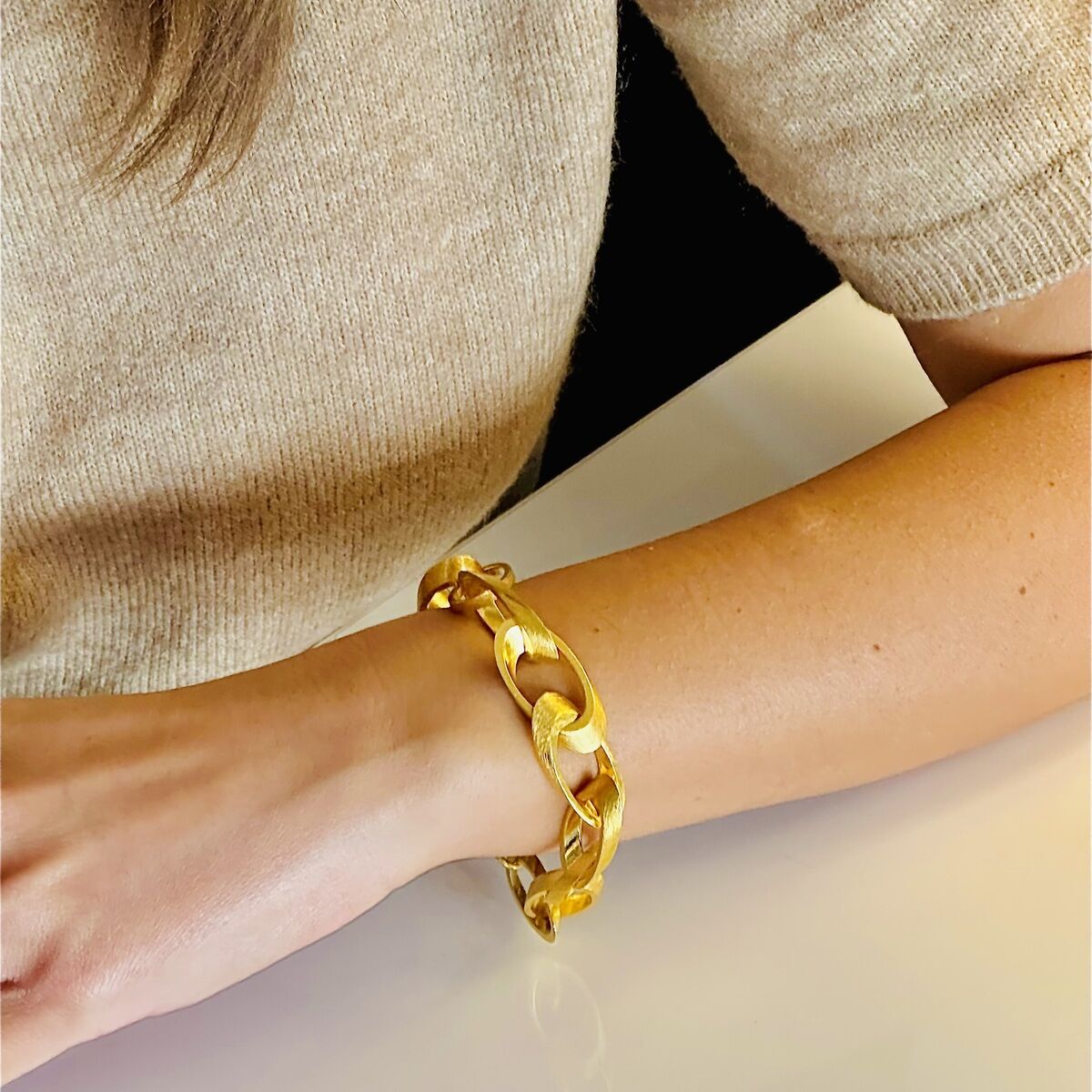 Eine Person trägt ein goldplattiertes Armband namens 'Jackie' aus Silber an ihrem Handgelenk. Das Armband hat große Glieder und liegt auf einem weißen Tisch. Der Ärmel des Oberteils der Person ist beige.
