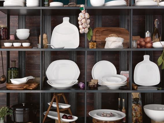 Regal mit weißen Keramiktellern, Schalen, Holzbrett, Knoblauch und Zwiebeln vor dunkler Wand
