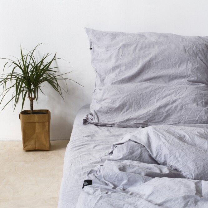 Eine gemütlich aussehende Bettwäsche in grauer Farbe auf einem Bett, daneben eine grüne Zimmerpflanze in einem braunen Topf. Die Wand im Hintergrund ist weiß.