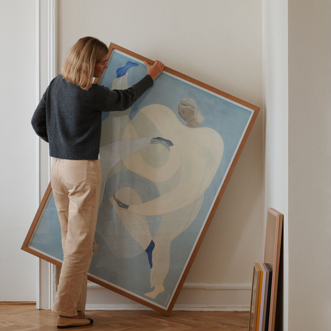 Eine Frau hängt ein großes Kunstwerk von Sofia Lind mit dem Titel 'Hold You - Blue' an eine weiße Wand. Das Bild zeigt eine abstrakte Figur in Blau- und Beigetönen. Weitere Bilder stehen an der Wand angelehnt.