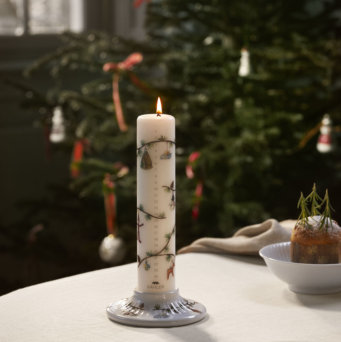 Eine brennende Adventskalenderkerze mit einem dekorativen Ständer steht auf einem gedeckten Tisch vor einem geschmückten Tannenbaum. Um die Kerze sind Zahlen und kleine weihnachtliche Motive aufgedruckt.