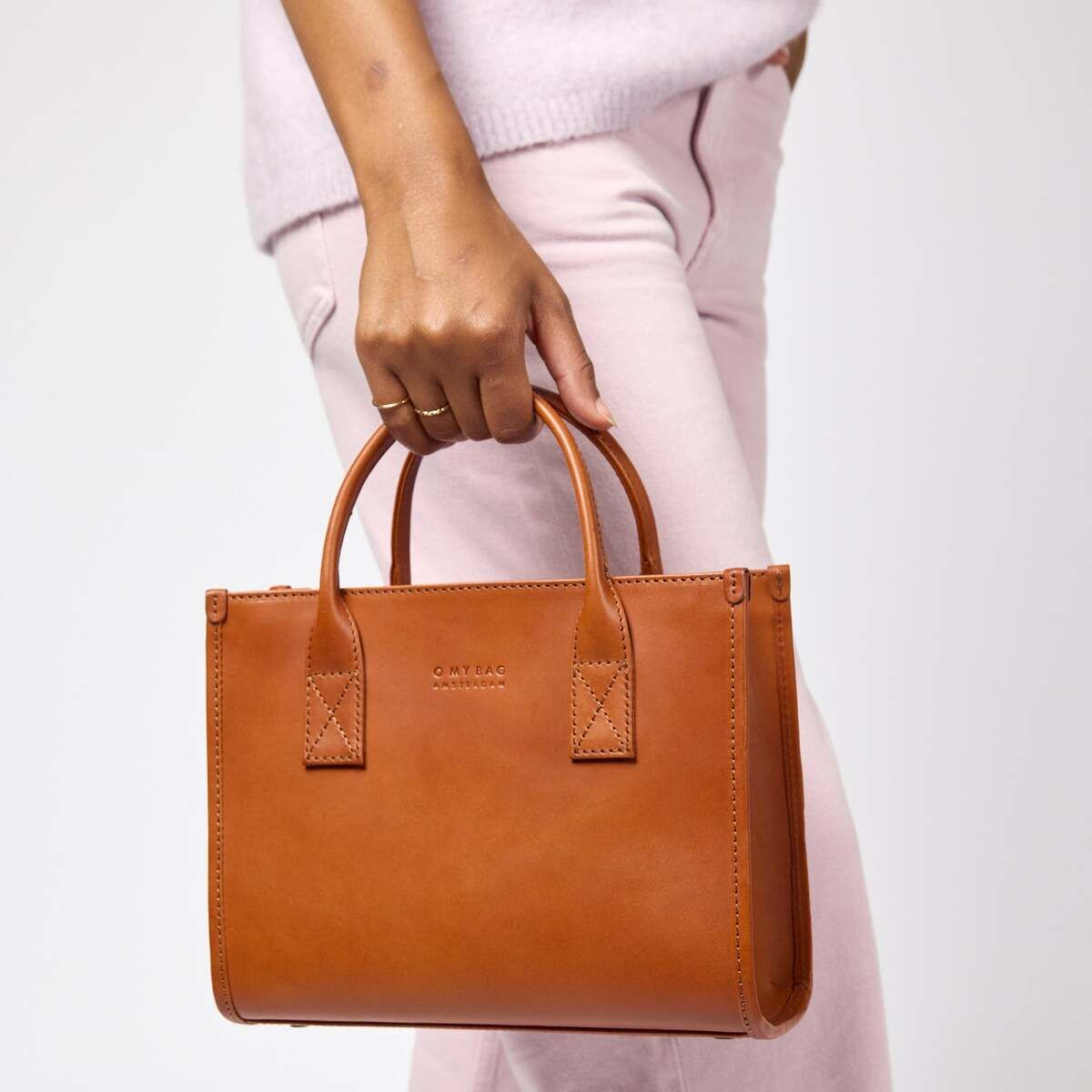 Nahaufnahme einer mittelgroßen, hellbraunen Ledertasche mit zwei Henkeln, die von einer Person in hellrosafarbener Kleidung getragen wird. Die Tasche hat ein minimalistisches, modernes Design und wirkt hochwertig verarbeitet. Das Foto eignet sich für Mode- und Accessoire-Präsentationen.