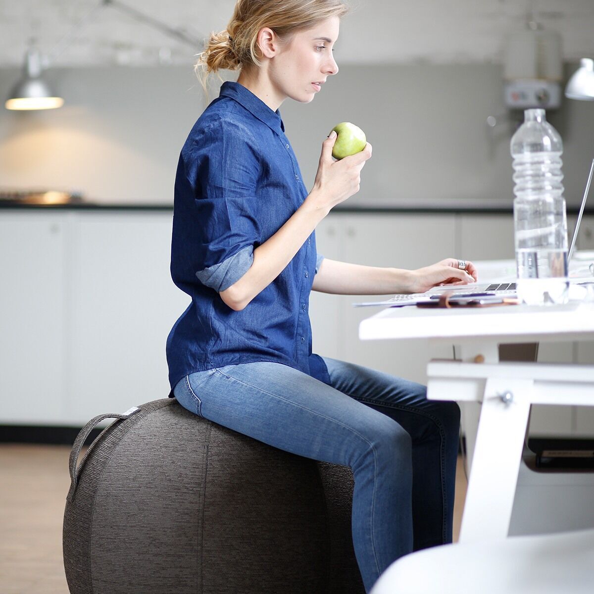 Eine Frau sitzt auf einem grauergonomischen Sitzball und hält einen grünen Apfel, während sie an einem weißen Tisch im Büro sitzt.