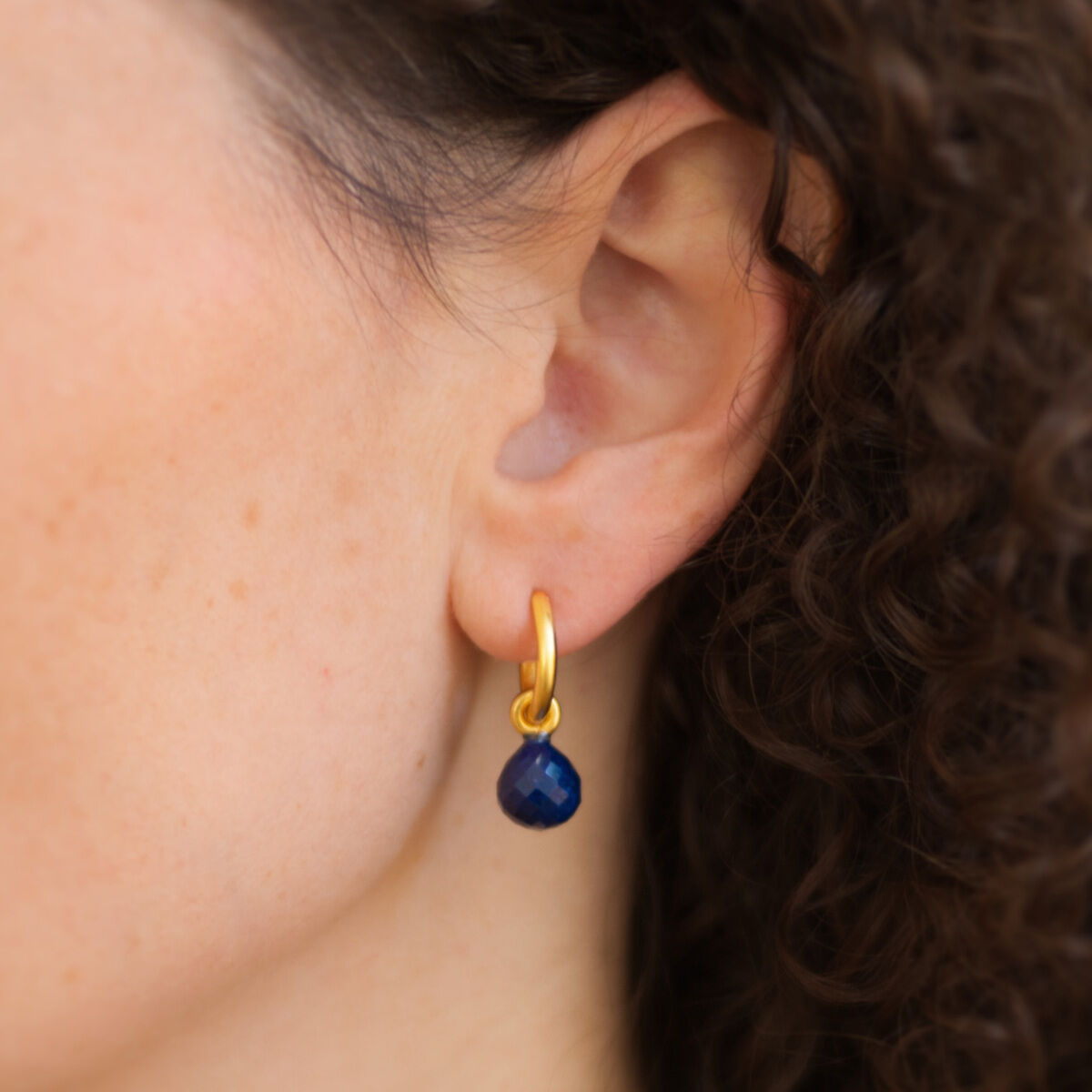 Detailaufnahme eines weiblichen Ohrs mit einer goldplattierten Creole von Dorle Pätzold Schmuck. Am Ohrring hängt ein facettierter dunkelblauer Lapislazuli-Tropfen. Das Schmuckstück ist elegant und edel, betont durch das dezente Design und hochwertige Materialien.