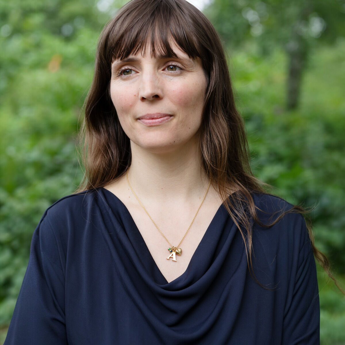 Frau mit braunen Haaren trägt eine vergoldete Halskette mit Buchstabenanhänger vor einer grünen, natürlichen Kulisse.