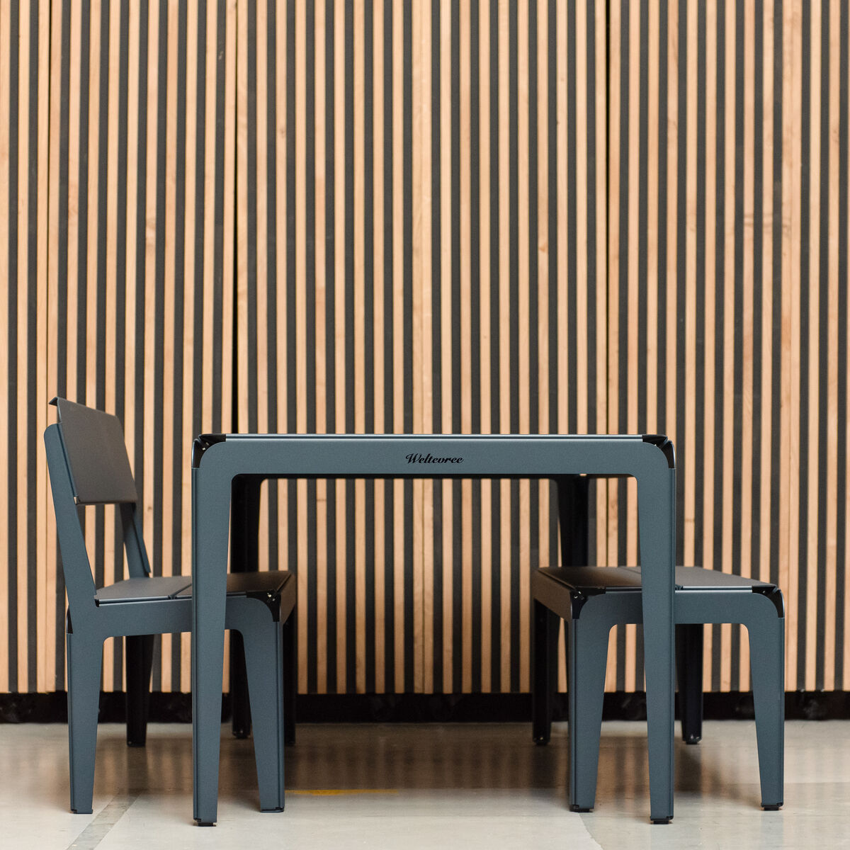 Eine moderne Bank mit Rückenlehne aus der Serie »Bended Bench«, platziert vor einem Hintergrund aus hölzernen vertikalen Streifen. Die Bank hat ein schlichtes und minimalistisches Design, ideal für den Innen- und Außenbereich.