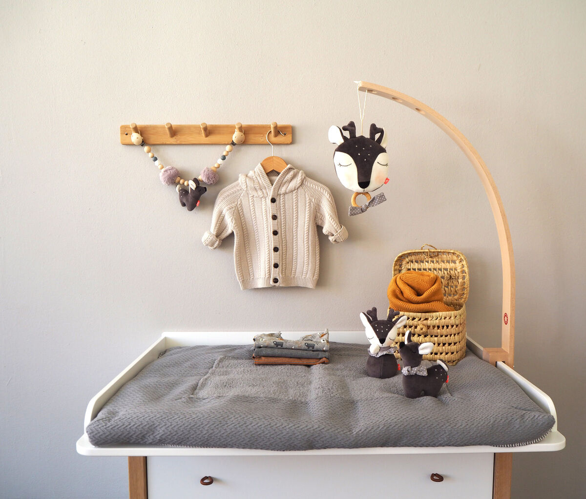 Ein mit einem Rehkitz und anderen Plüschtieren dekoriertes Babyzimmer mit einer Spieluhr an einem Holzgestell über einer Wickelkommode. An der Wand hängt ein Strickpullover neben einer Schnullerkette mit einem Plüsch-Elch.