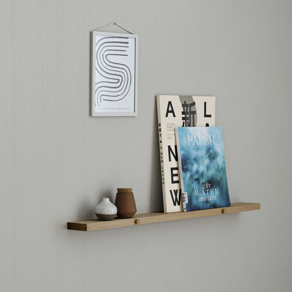 Ein schlichtes Regal aus Eichenholz ist an einer hellgrauen Wand montiert. Auf dem Regal stehen verschiedene Dekorationsgegenstände und Bücher. Darunter eine kleine braune Vase, weiße Keramik und eindrucksvolle Zeitschriften.