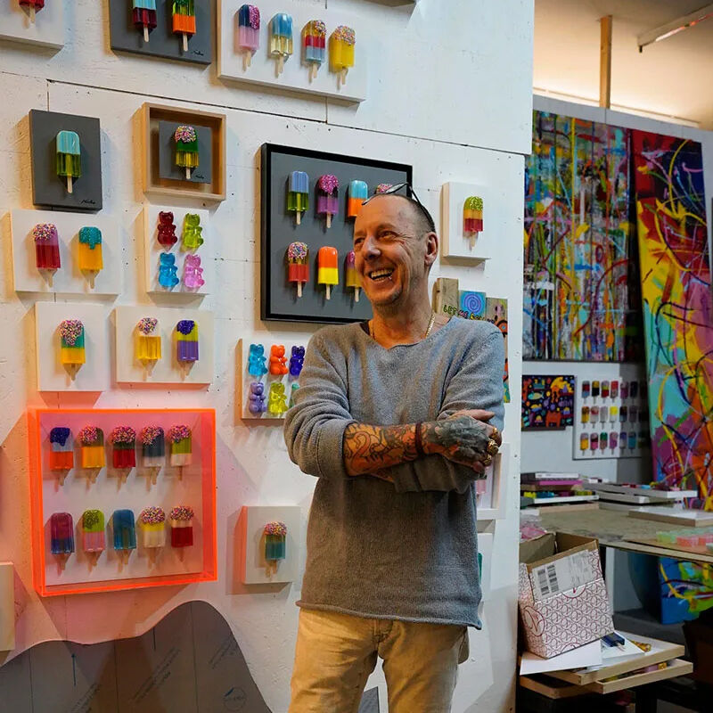 In einem modernen Atelier steht ein lächelnder Mann vor einer Wand, an der zahlreiche farbige, gerahmte Kunstdrucke von Eis am Stiel in verschiedenen Formen und Farben hängen. Im Hintergrund sind weitere Kunstwerke und Materialien sichtbar, die den kreativen Raum betonen.
