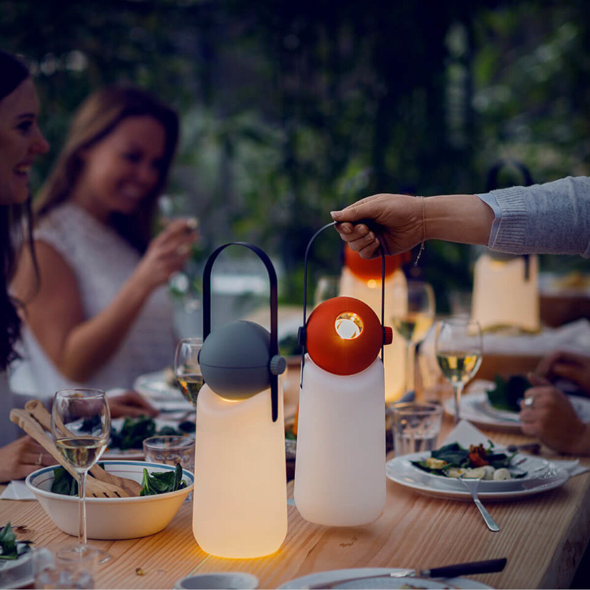 Draußen sitzen mehrere Personen an einem gedeckten Tisch. Auf dem Tisch stehen leuchtende, tragbare »Guidelight« Lampen in Weiß und Grau mit orangefarbenen Details. Die Beleuchtung schafft eine gemütliche Atmosphäre.