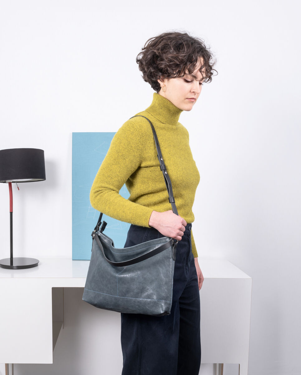 Eine Frau in einem senfgelben Rollkragenpullover und dunkelblauer Hose trägt eine graue Leder-Schultertasche »Nabuko«. Im Hintergrund sind ein weißer Tisch, ein schwarz-roter Tischlampen und ein blaues Bild zu sehen.