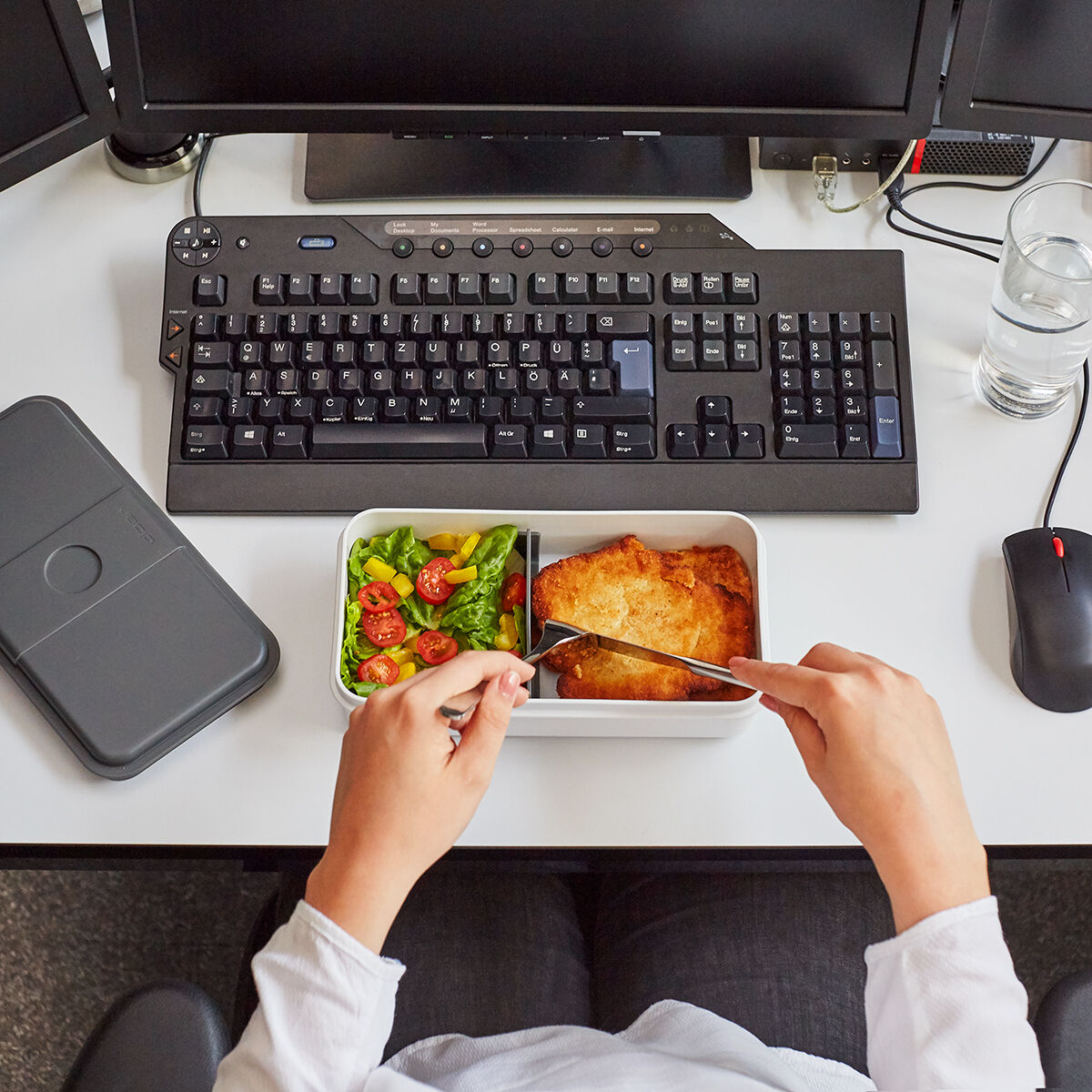 Eine Person sitzt an einem Schreibtisch mit mehreren Computerbildschirmen und bereitet ein Mittagessen in einer Lunchbox vor. Die Lunchbox enthält verschiedene gesunde Lebensmittel, darunter Kirsch-Tomaten und grüner Salat. Auf dem Schreibtisch befinden sich eine Tastatur, eine Maus, ein Glas Wasser und eine weitere geschlossene Lunchbox.