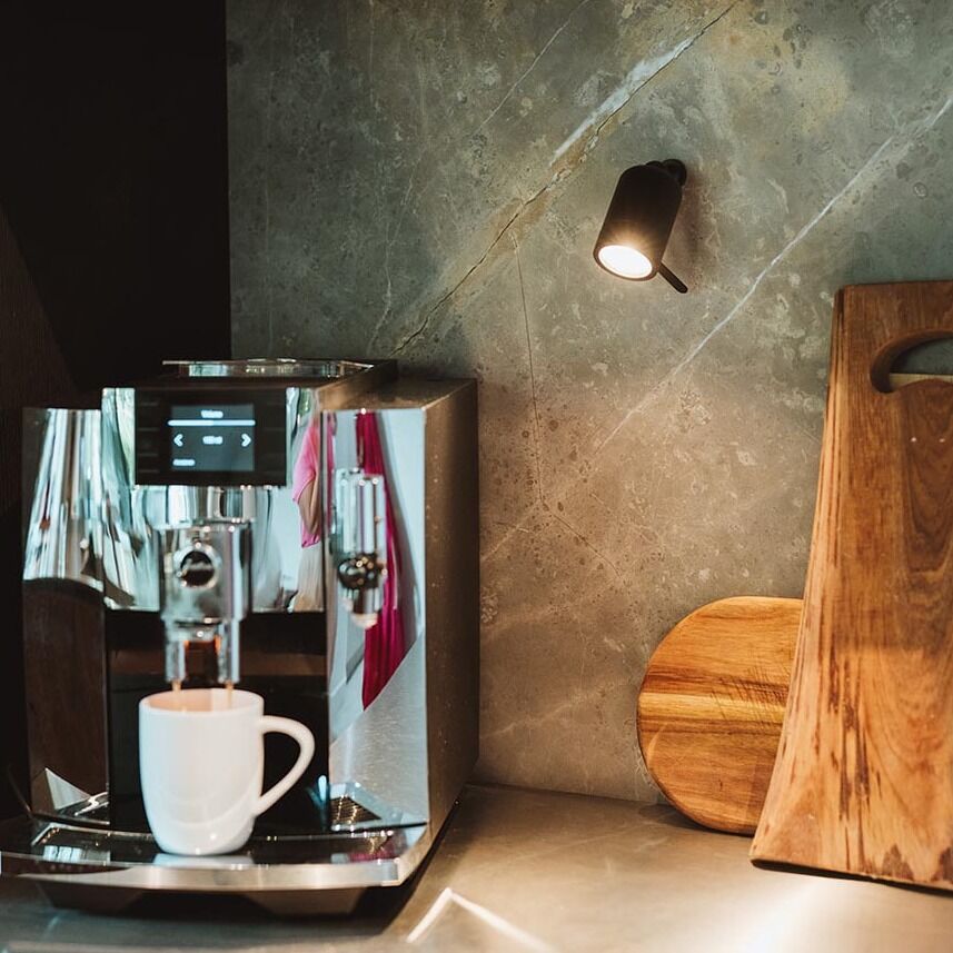 Eine Küche mit einer Edelstahl-Kaffeemaschine auf der Arbeitsfläche, daneben Holzschneidebretter und eine Wandleuchte, die eine Tasse beleuchtet. Im Hintergrund ist eine Wand aus Marmor und ein Bücherregal zu sehen.