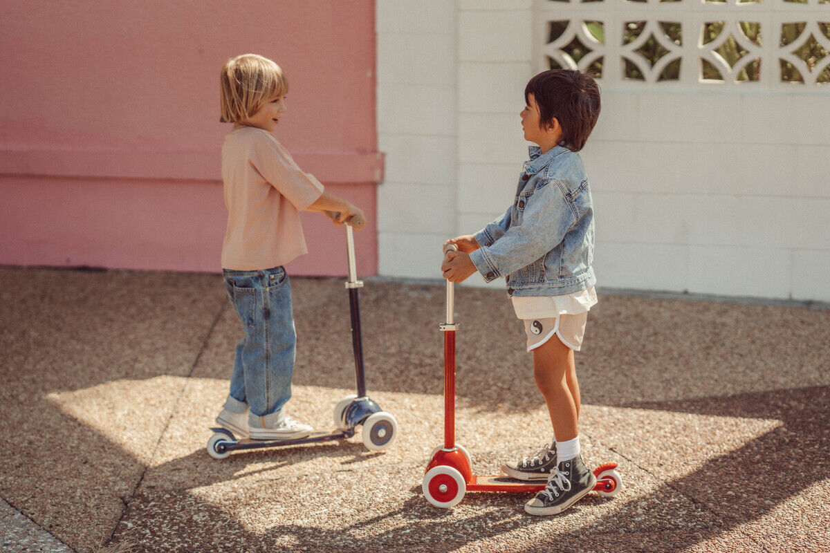 Zwei Kinder fahren auf Scootern im Freien. Ein Kind trägt eine rosa Bluse und blaue Jeans, das andere Kind eine Jeansjacke und weiße Shorts. Der Hintergrund zeigt eine Mauer in Rosa und Weiß.