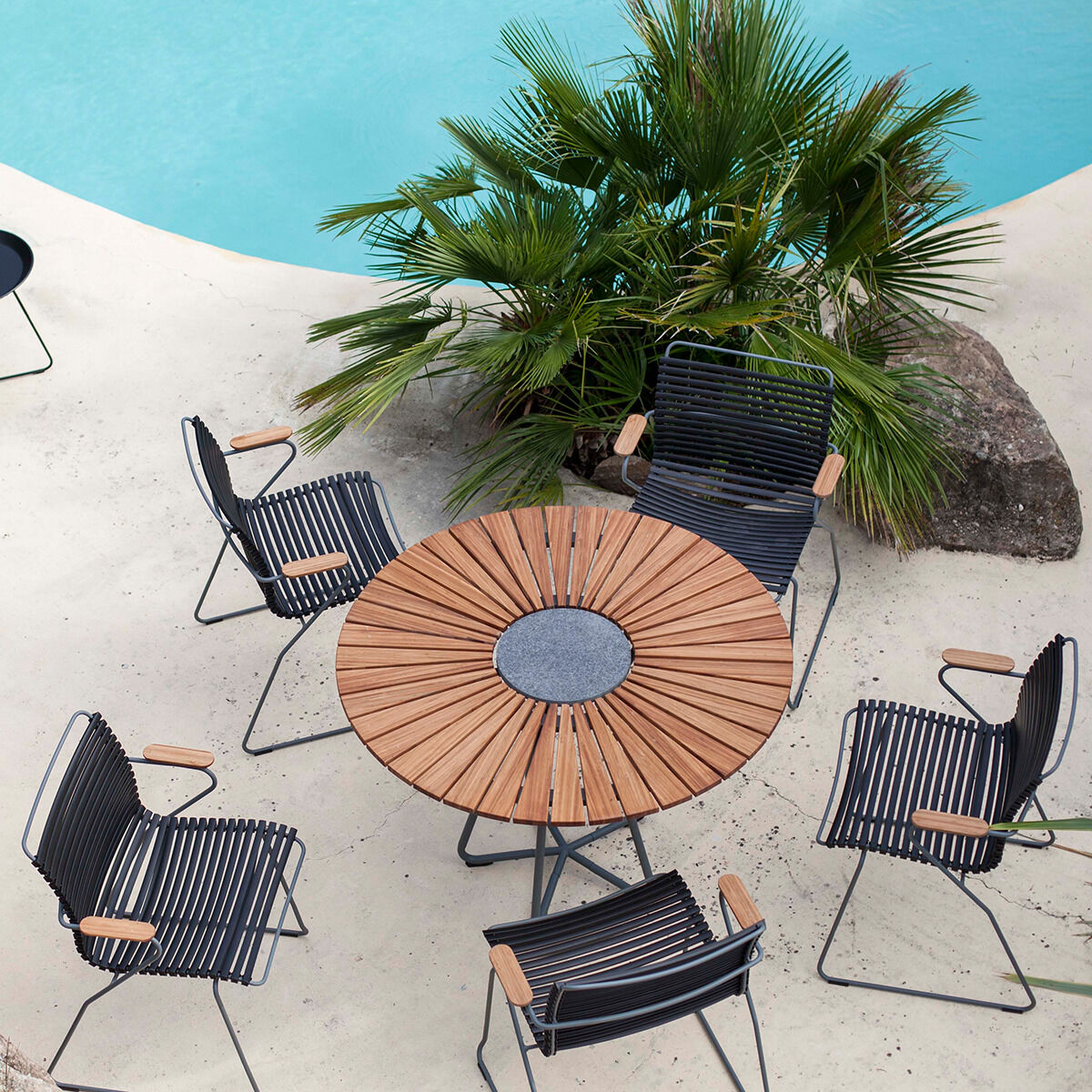 Gartentisch »CIRCLE«: Ein runder Holztisch mit Metallgestell, umgeben von fünf Gartenstühlen mit Holzarmlehnen und Metallrahmen. Im Hintergrund sind zahlreiche Palmen und ein Pool zu sehen, die eine erholsame Outdoor-Ambiance schaffen.