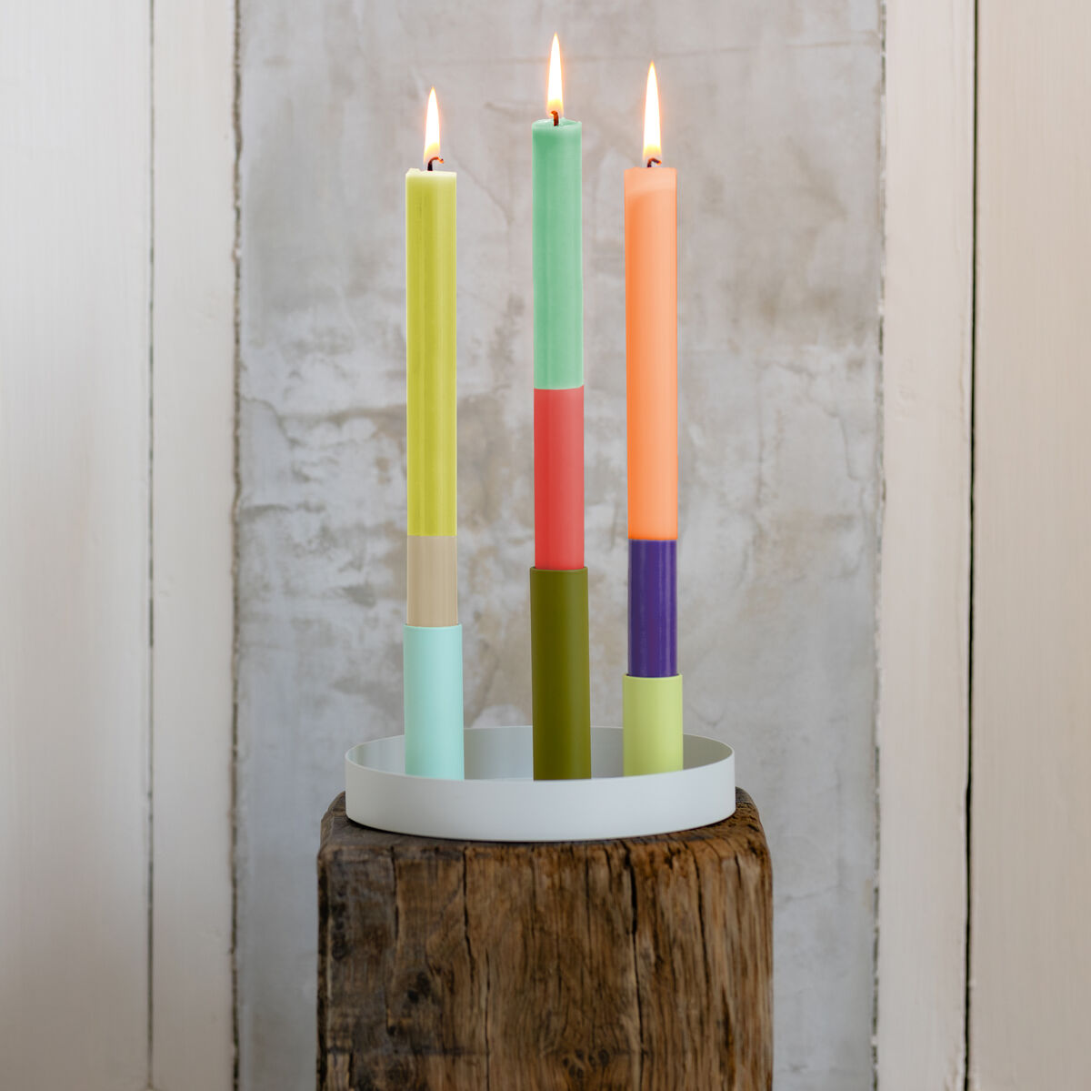 Ein Set aus bunten, aufrechten Stabkerzen in verschiedenen Farben (grün, rosa, orange, gelb) auf einem weißen Kerzenteller, auf einem rustikalen Holzpodest vor einer weißen und grauen Wand platziert.