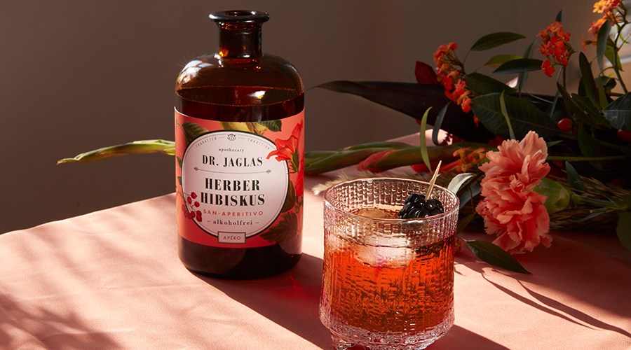 Eine Flasche Dr. Jaglas Herber Hibiskus steht neben einem Glas Hibiskusgetränk, garniert mit Kirschen, umgeben von Blumen auf einer rosa Tischdecke.