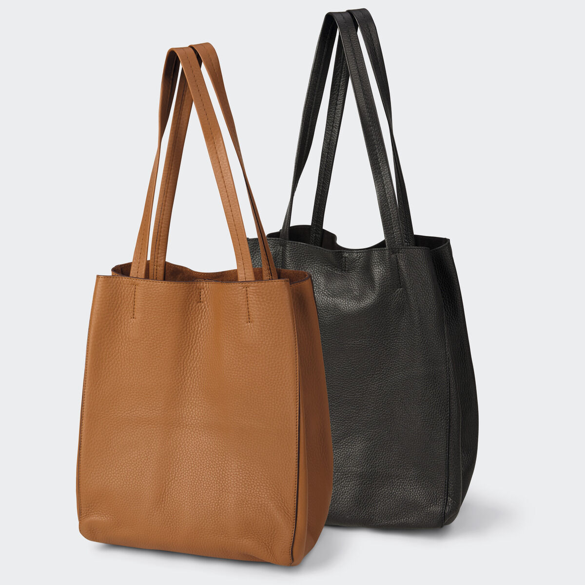 Zwei elegante, schlichte Ledertaschen der Marke Guerer & Guerer: vorn eine Tasche in Cognac, dahinter eine in Schwarzbraun. Beide haben lange Henkel, weiche, strukturierte Oberfläche und ein reduziertes, modernes Design. Freistehend vor hellem Hintergrund.