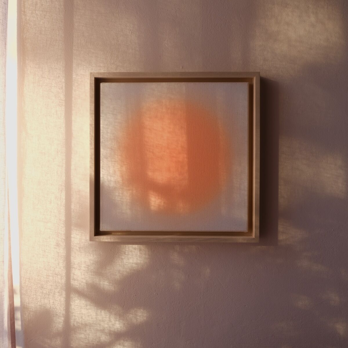 Quadratisches Kunstwerk in hellem Holzrahmen an einer beige-rosafarbenen Wand. Im Zentrum des Bildes befindet sich ein weicher, orangefarbener Kreis vor einem dezent hellen Hintergrund. Sonnenlicht und Schatten aus einem Fenster erzeugen atmosphärische Lichtspiele.