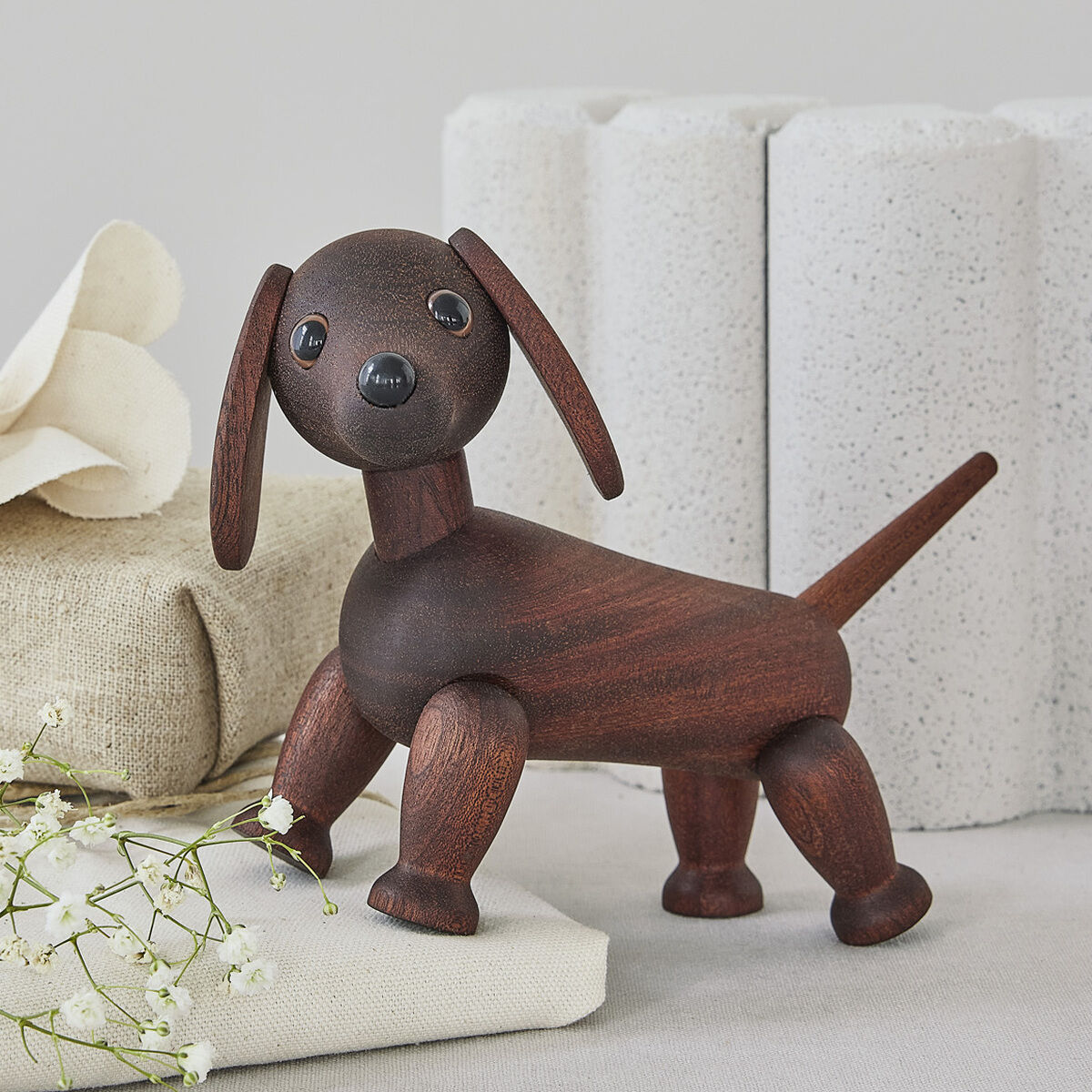 Die Holzfigur »Dackel Quick« von Spring Copenhagen zeigt einen stilisierten, dunkelbraunen Dackel aus glattem Holz. Sie steht auf hellen Stoffen und neben Geschenkpaketen mit weißen Blüten, vor hellgrauem Hintergrund mit rechteckigen Dekoelementen.