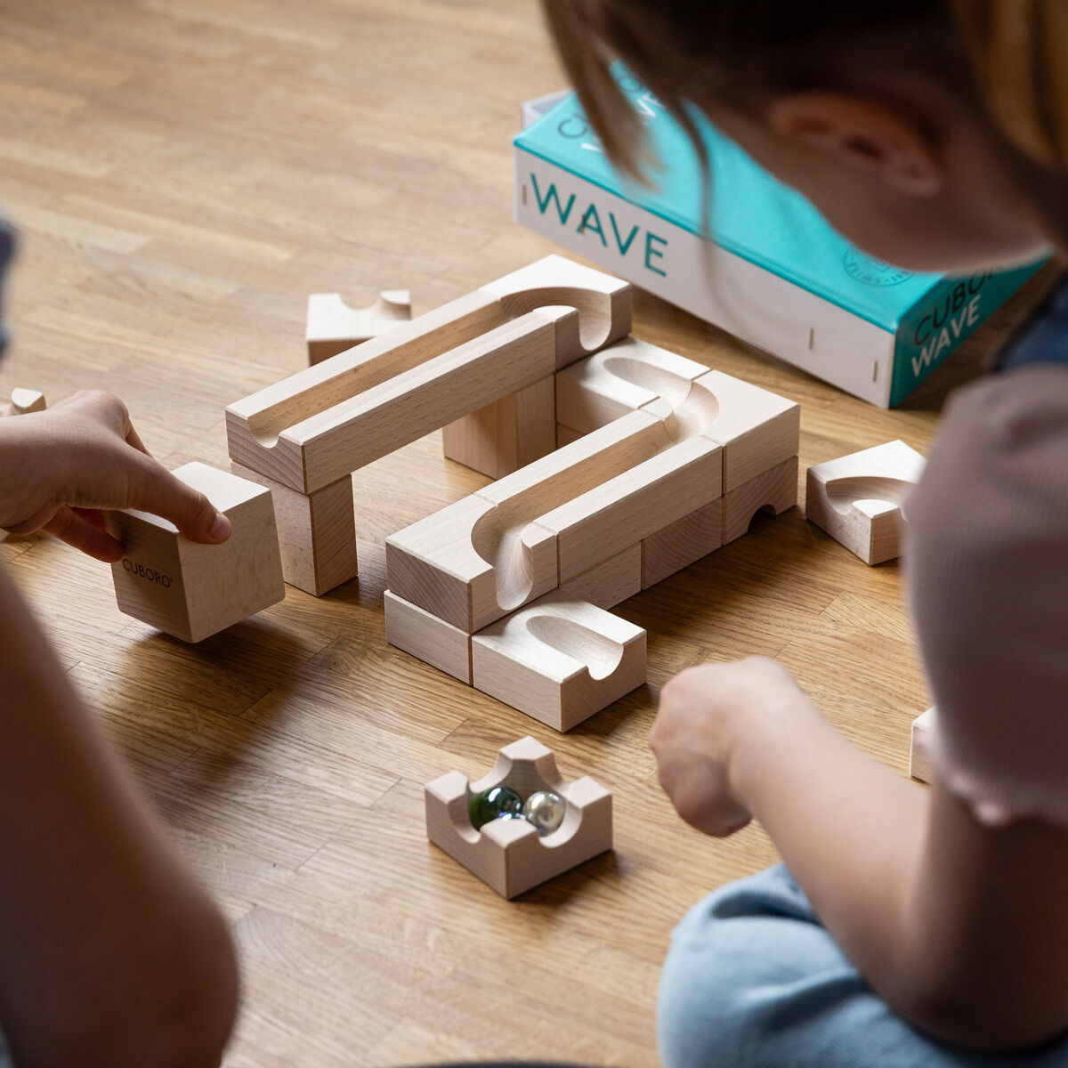 Zwei Kinder spielen auf dem Boden mit einem Set von Holzbausteinen, um eine Murmelbahn zu bauen. Die Szene zeigt kreative Aktivitäten mit Holzspielzeug.