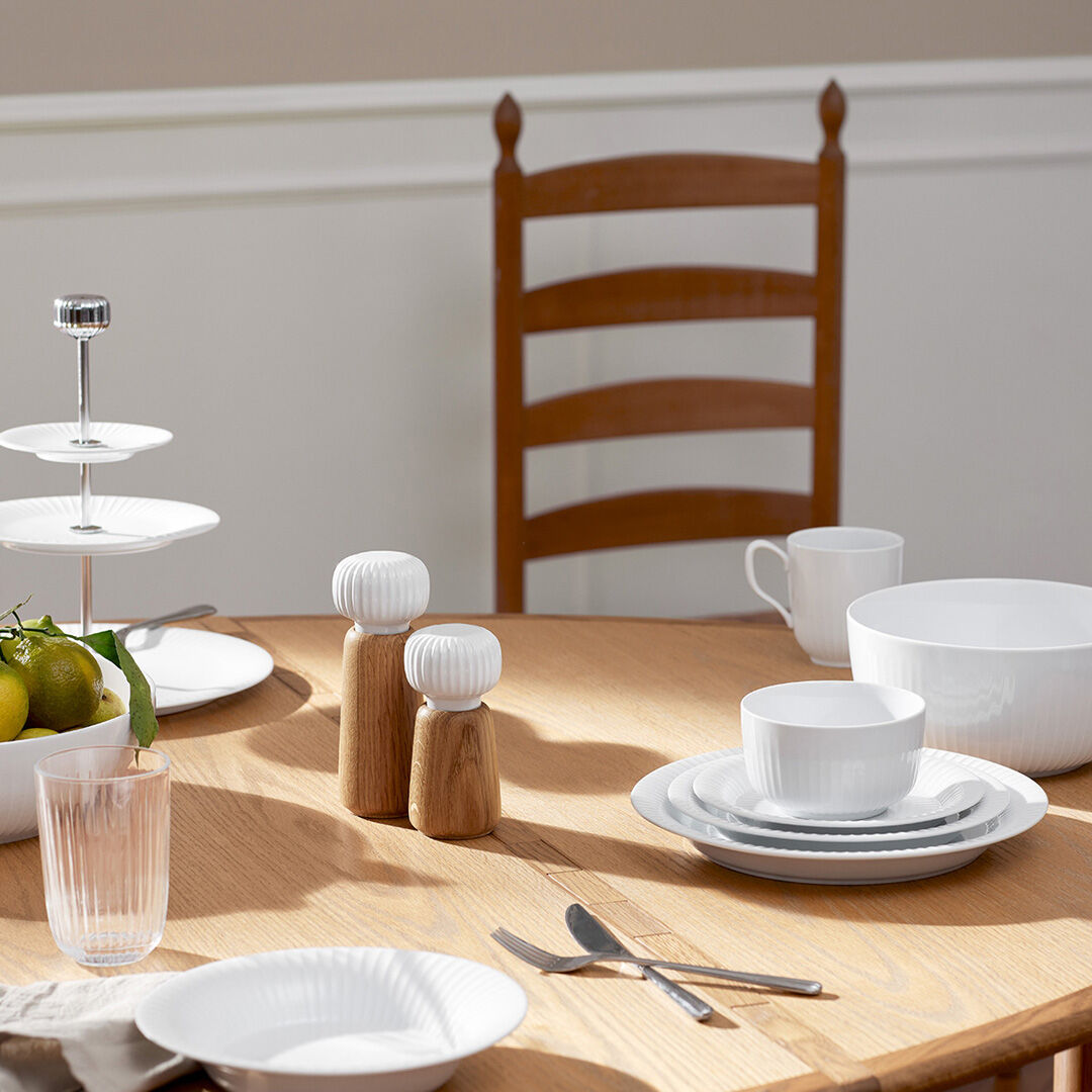 Auf einem hellen Holztisch steht eine skandinavische Gewürzmühle der Serie »Hammershøi«, flankiert von weiterem weißen Geschirr, einer Etagere mit Obst und einem Wasserglas. Im Hintergrund ist ein Holzstuhl zu sehen. Die Szene vermittelt ein helles, modernes und einladendes Ambiente für den gedeckten Esstisch.