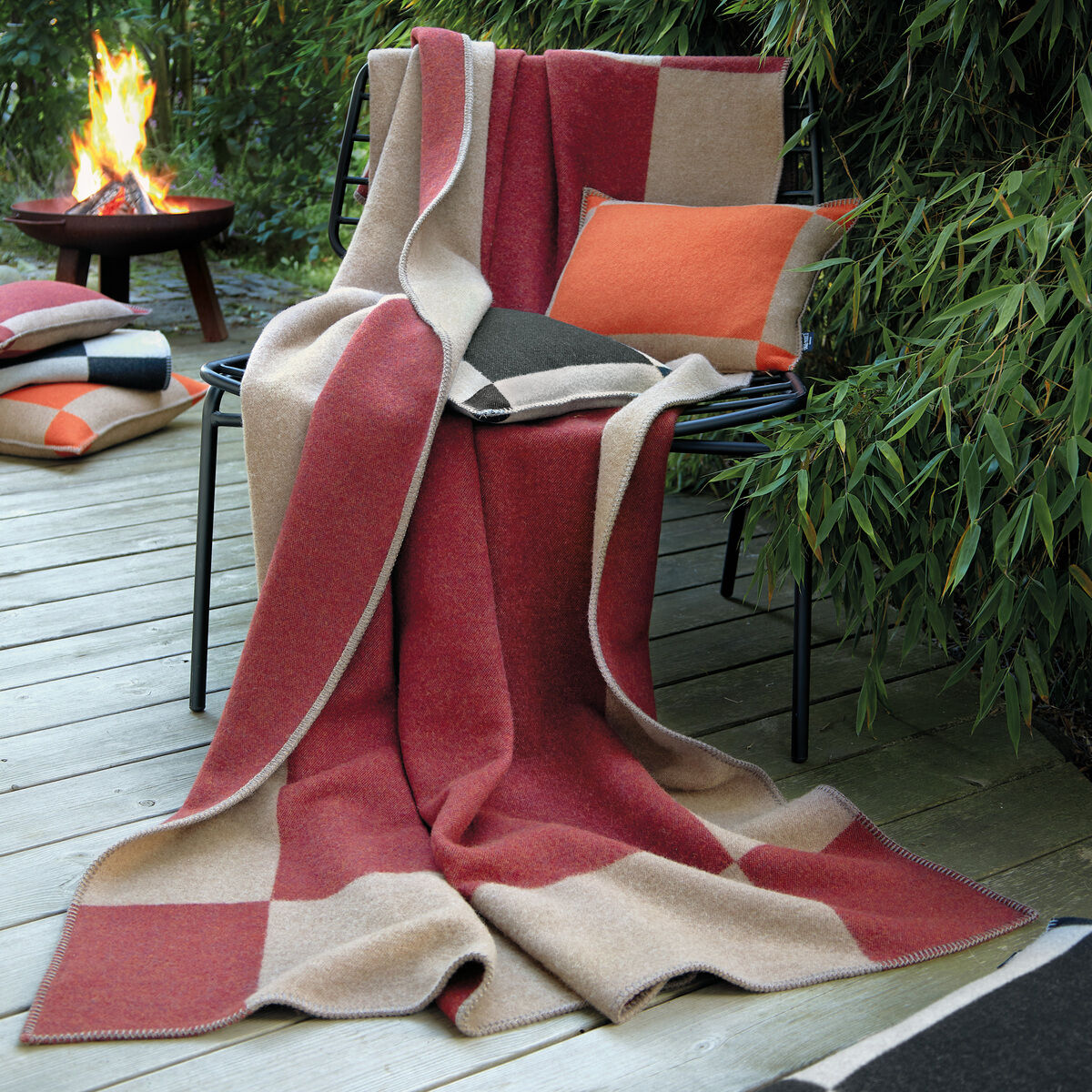 Gemütliche Decke »Manchester« in Rot, Beige und Schwarz auf einem Stuhl. Im Hintergrund ein Feuer und Bambussträucher. Ein orangefarbenes Kissen auf dem Stuhl und ein weiteres Kissen am Boden ergänzen die Szene. Perfekt für drinnen und draußen, insbesondere in der kühleren Jahreszeit.