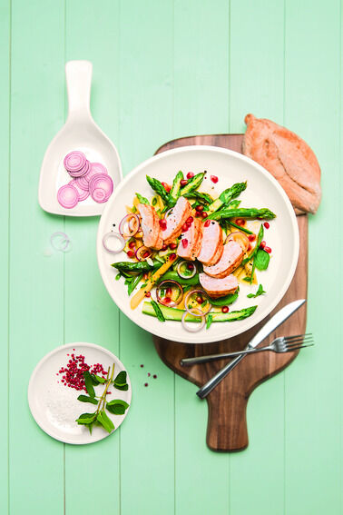 Das Bild zeigt ein Holzschneide- und Servierbrett mit einem darauf liegenden Teller, der ein Gericht aus Fleisch, Spargel, Zwiebeln und Granatapfelkernen enthält. Daneben liegen eine Scheibe Brot, ein kleines Tellerchen mit Salz und rosa Beeren und ein Porzellangeschirr mit in Scheiben geschnittenen Zwiebeln. Der Hintergrund besteht aus grünen Holzdielen.