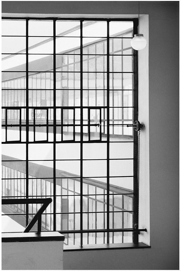 Schwarzweißfoto von Klaus Kinold unter dem Titel 'Walter Gropius, Bauhaus Dessau, Blick vom Treppenhaus nach außen, 1992'. Die Aufnahme zeigt ein modernes, geometrisches Fenstergitter, das den Blick nach außen auf ein weiteres Bauhaus-Gebäude ermöglicht.