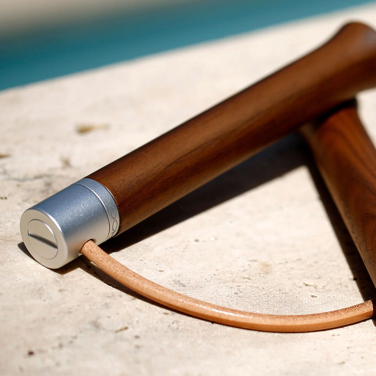 Detailaufnahme des Springseils »ROTATOR« von HOCK Design: Zwei edle, geschwungene Handgriffe aus amerikanischem Nussbaum mit feinen Aluminium-Enden und einem naturfarbenen Lederseil, stilvoll platziert auf einer hellen Steinoberfläche, im Hintergrund unscharf Blauaquarell.