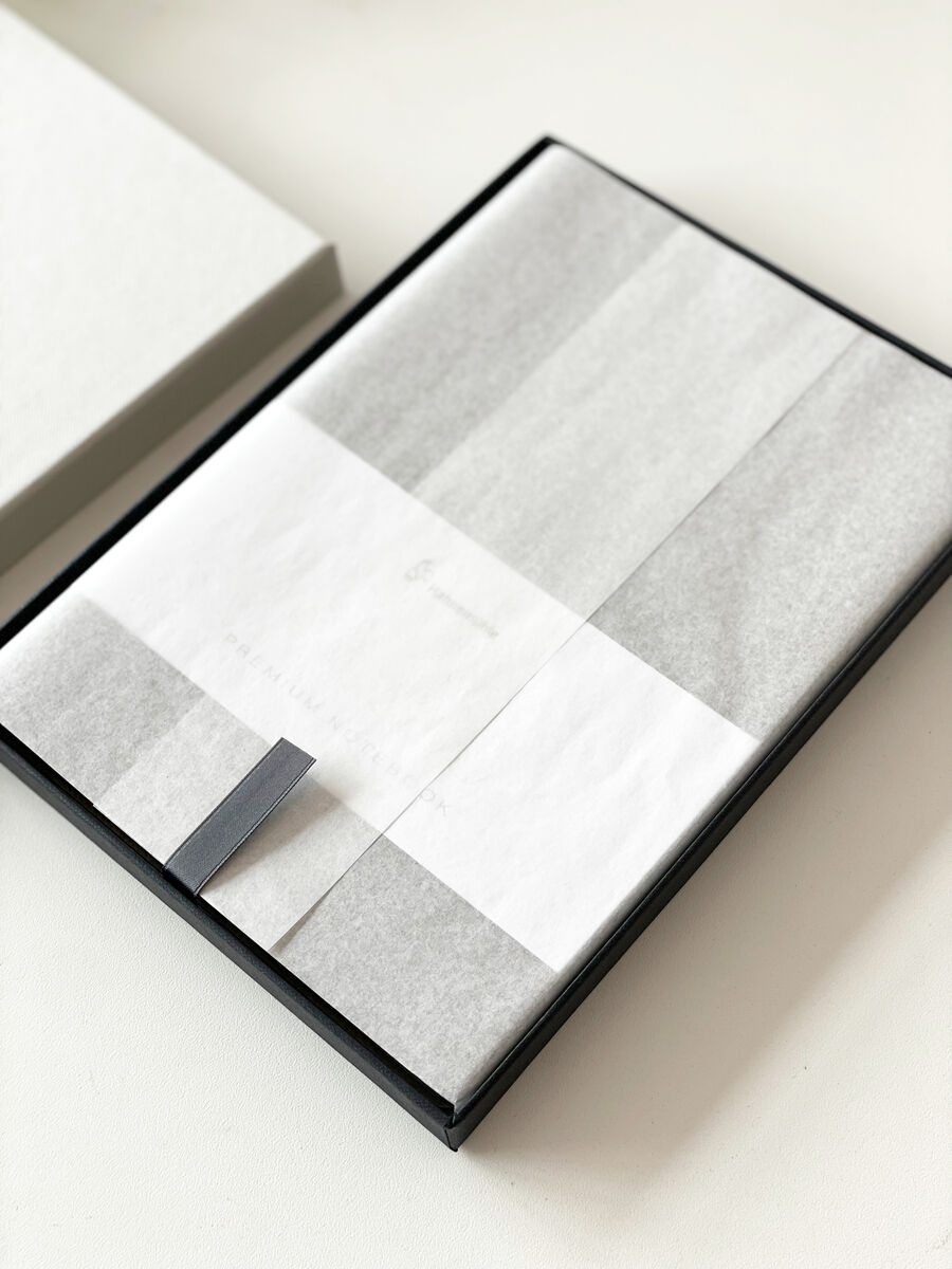 Ein stilvolles Notizbuch im DIN A5-Format, eingehüllt in graues und weißes Papier, präsentiert in einer eleganten schwarzen Box. Perfekt für Notizen, Planungen oder als Geschenk.