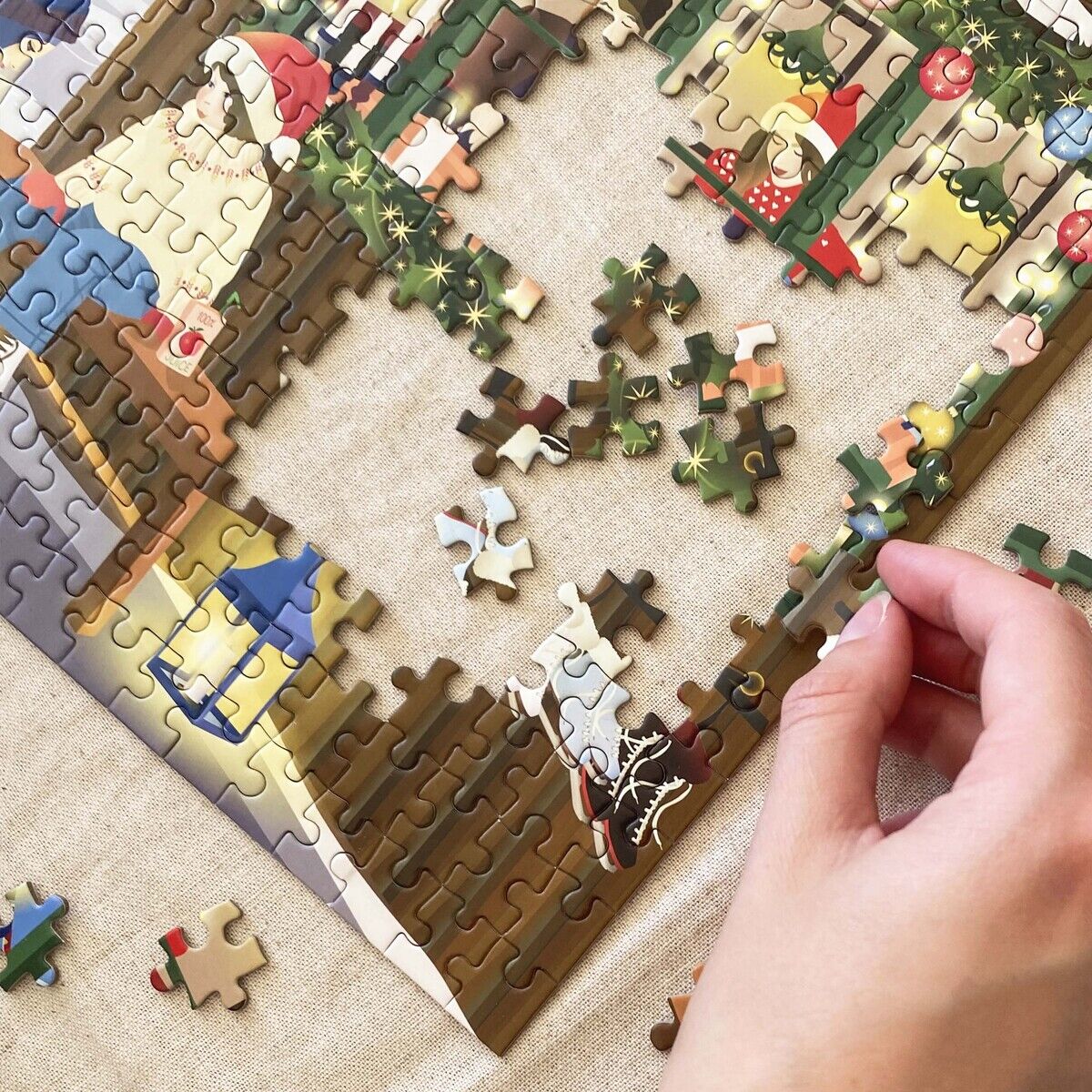 Nahaufnahme eines teilweise fertiggestellten Puzzles mit 1000 Teilen auf einem beigen Untergrund. Ein Teilmotiv zeigt einen Kaffee-Laden mit skandinavischer Dekoration, weihnachtlicher Girlande, Lampions und Figuren. Eine Hand setzt ein Teil ein. Marke: ViSSEVASSE.